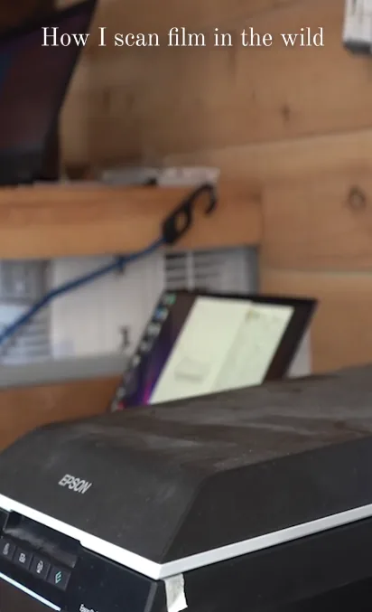 An Epson flatbed scanner on a wooden shelf in a cabin, with a laptop showing scanning software behind it and the title "How I scan film in the wild" across the top.