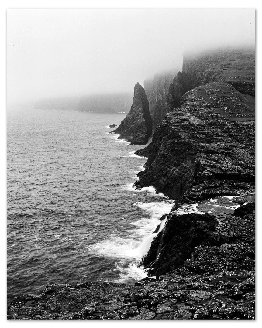 Black and white photo of sea cliffs in fog in the Faroe Islands: a pointed sea stack offshore, waves breaking on dark rock, and a stream falling off the cliff edge into the sea.